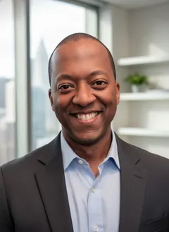 Carlus Henry smiling, wearing a gray suit jacket and open-collar light blue shirt, in a modern office with a city skyline visible through the windows behind him.